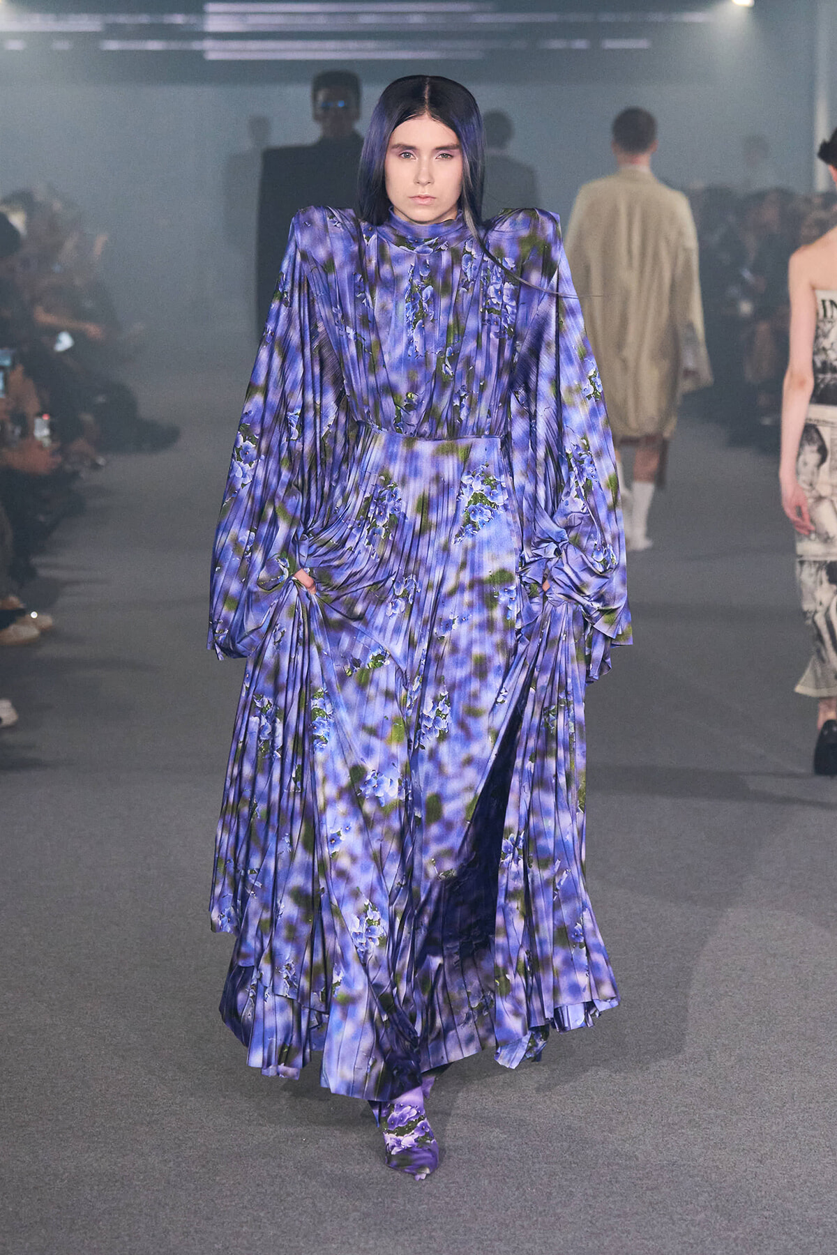 Model walks a fashion runway wearing a voluminous purple floral gown with structured shoulders and long draped sleeves.