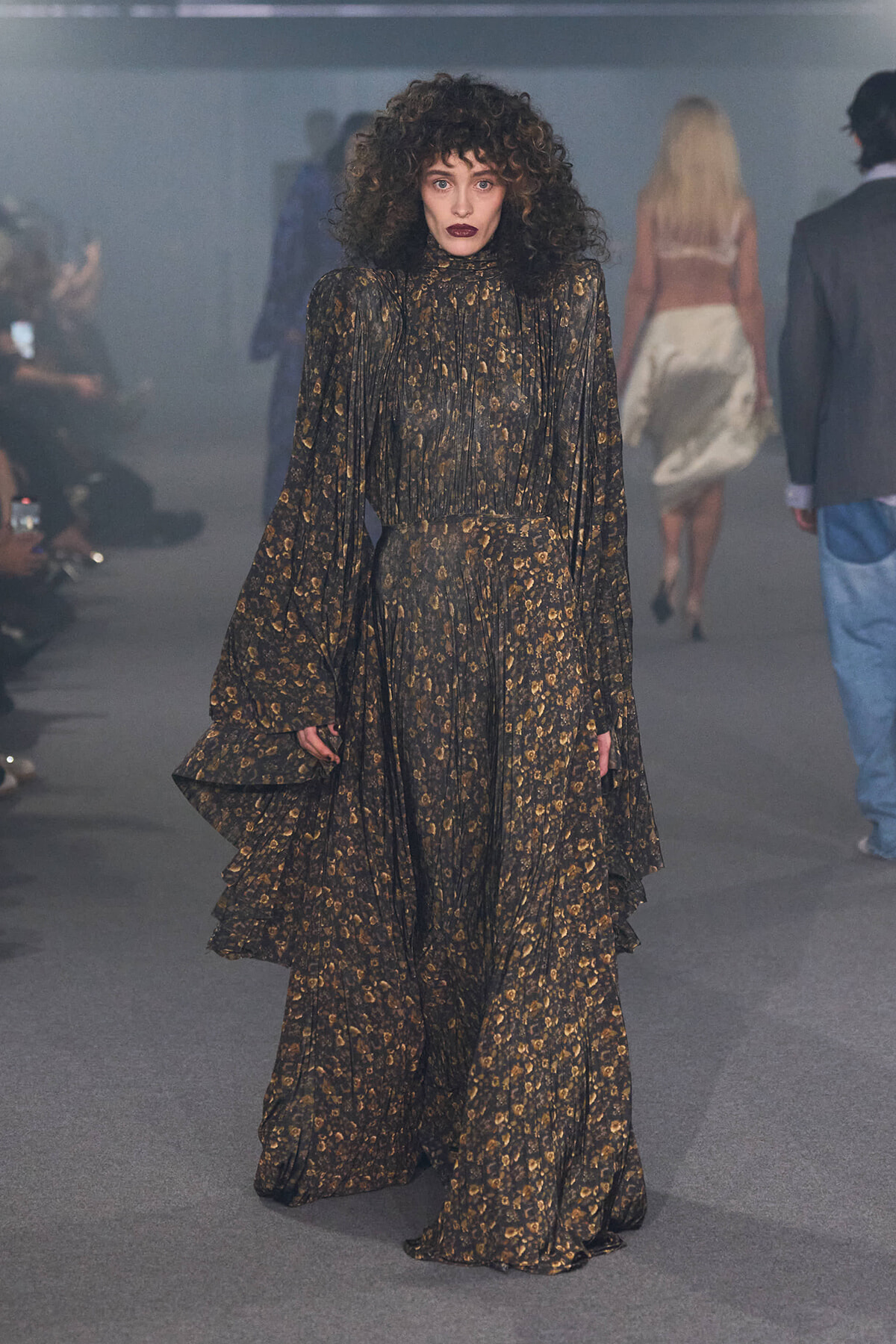 Model walks runway in a long, dark floral gown with layered ruffles and sheer sleeves.