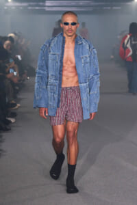 Male model on a runway wearing an oversized denim jacket, red plaid shorts, sunglasses, and no shirt, showcasing a casual denim look.