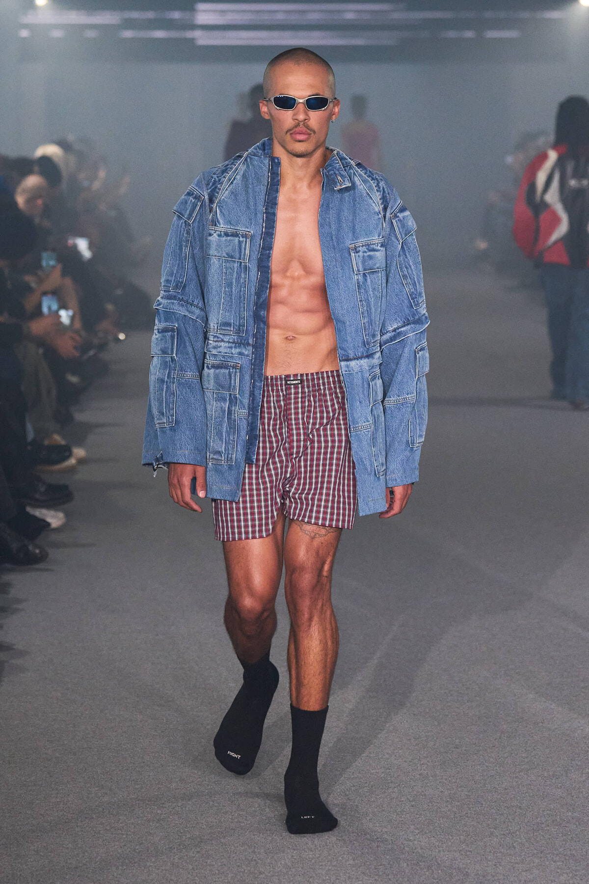 Male model on a runway wearing an oversized denim jacket, red plaid shorts, sunglasses, and no shirt, showcasing a casual denim look.