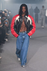 Male model on a fashion runway wearing a red and black leather jacket with white panels, open at the chest, and baggy blue jeans.