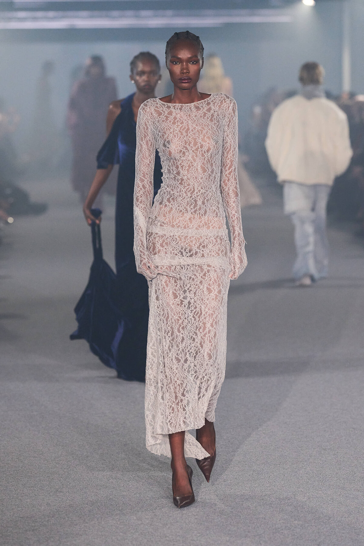Model walking the runway in a sheer pink lace gown with long sleeves and nude underlay, brown pointed heels.