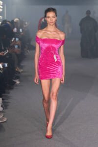 Model walking the runway in a bright pink off-the-shoulder velvet mini dress with a white tag hanging from the waist.