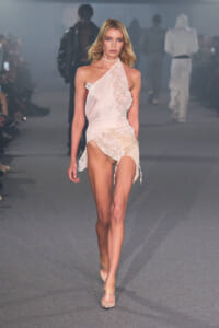 Model walking a runway in a pale pink lace wrap dress with an asymmetric one-shoulder neckline and nude heels