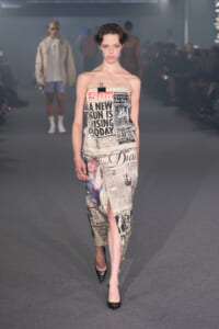 Model walks a runway wearing a strapless dress covered in newspaper print.