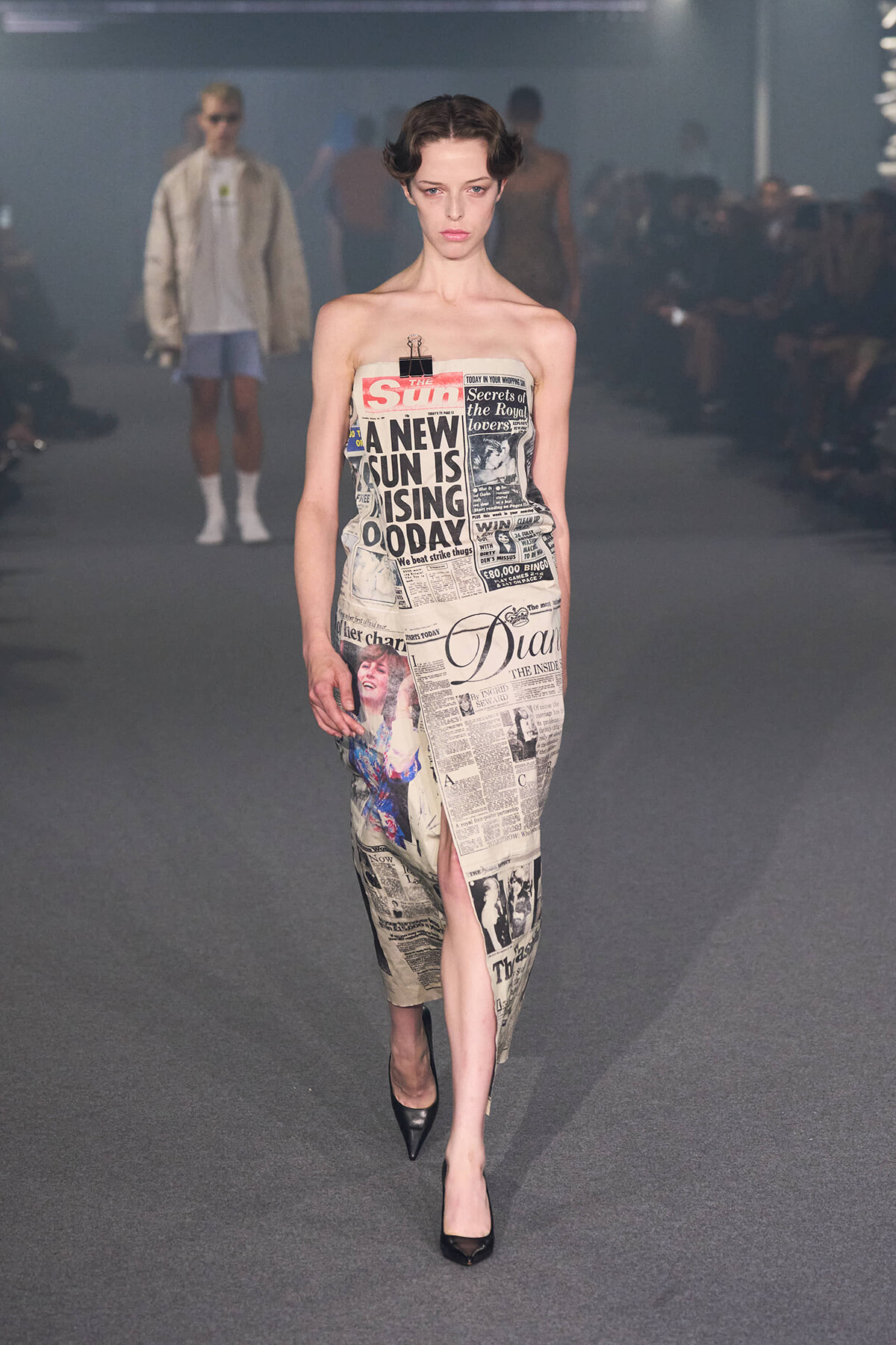 Model walks a runway wearing a strapless dress covered in newspaper print.