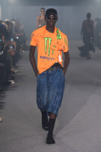 Male model walks a fashion show wearing an orange Monster T-shirt over a black undershirt, denim cargo shorts, and black socks with a cap and sunglasses.