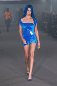 Model walking runway in a royal blue off-shoulder velvet mini dress with a white tag hanging from the chest, matching blue heels, audience blurred in background.