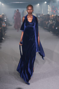 Model walking the runway in a floor-length navy velvet gown with a draped scarf-like detail and trailing hem.