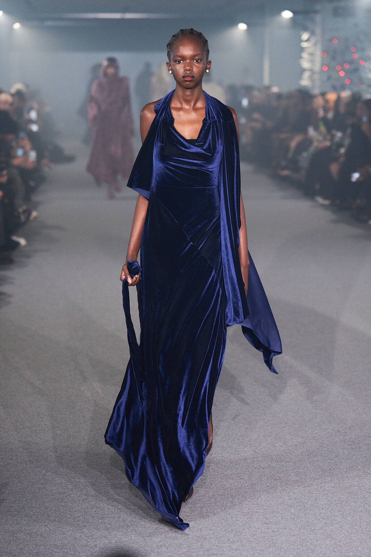 Model walking the runway in a floor-length navy velvet gown with a draped scarf-like detail and trailing hem.