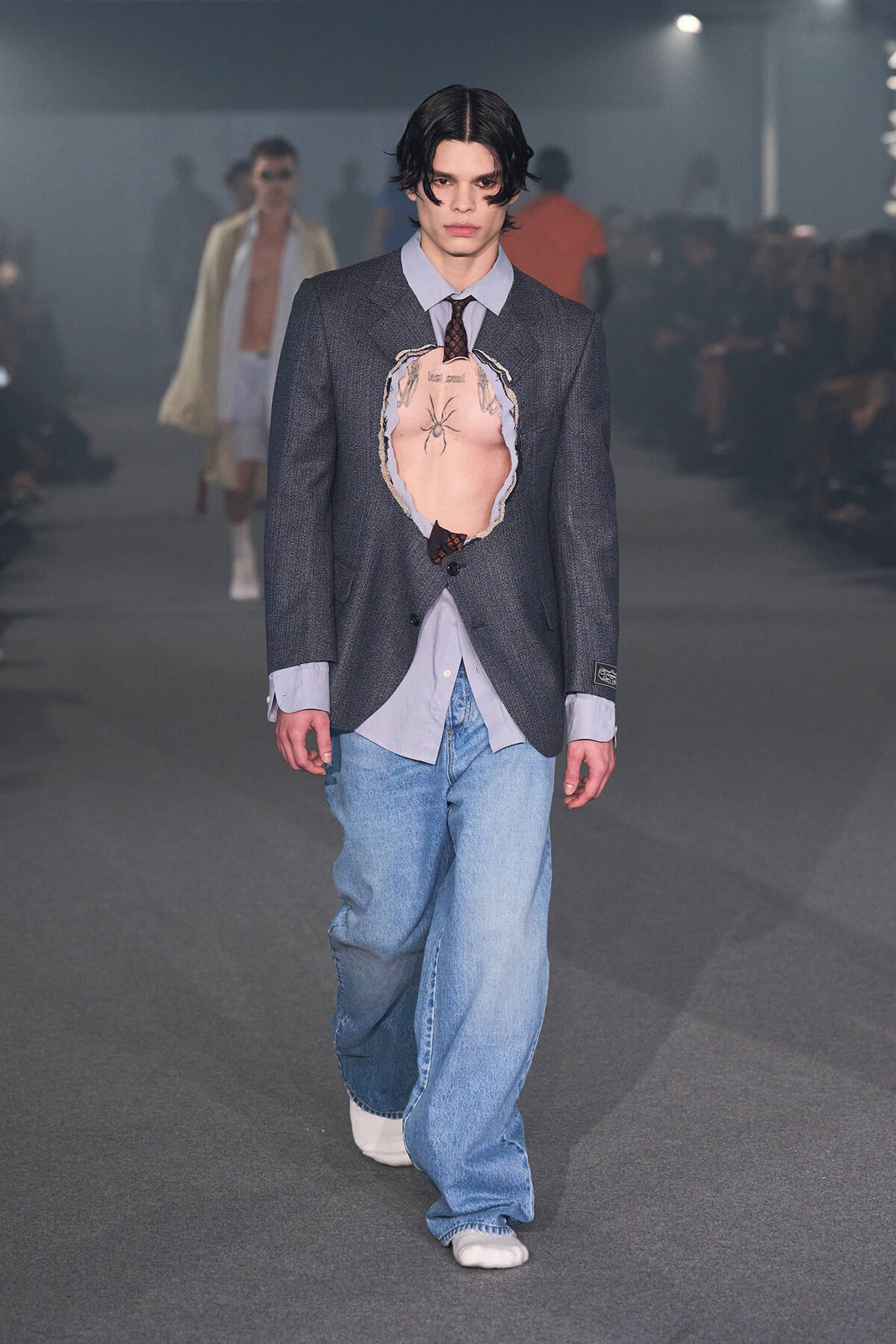 Male model on a runway wearing a dark blazer with a large chest cutout revealing a torso tattoo, paired with a light blue shirt, loose jeans, and white shoes; fashion show setting in background.