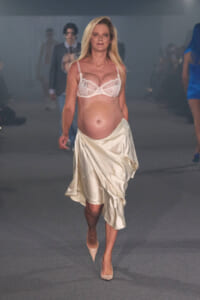 Pregnant model walking a fashion runway in a white lace bra and cream satin skirt with nude heels, blurred audience in the background.