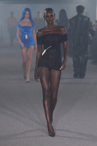 Dark-skinned runway model in a black off-shoulder mini dress with a white tag, walking toward the camera in a fashion show.