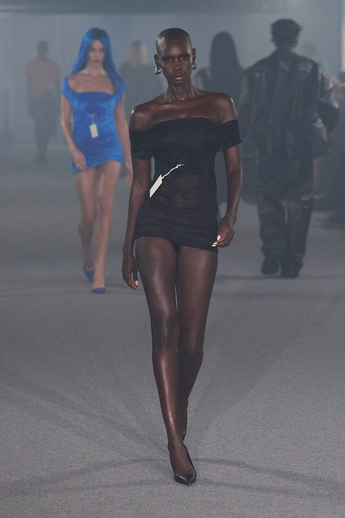 Dark-skinned runway model in a black off-shoulder mini dress with a white tag, walking toward the camera in a fashion show.