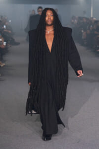 Model walks a runway in a loose black suit with extreme long dreadlocks and a cross necklace, minimal makeup.