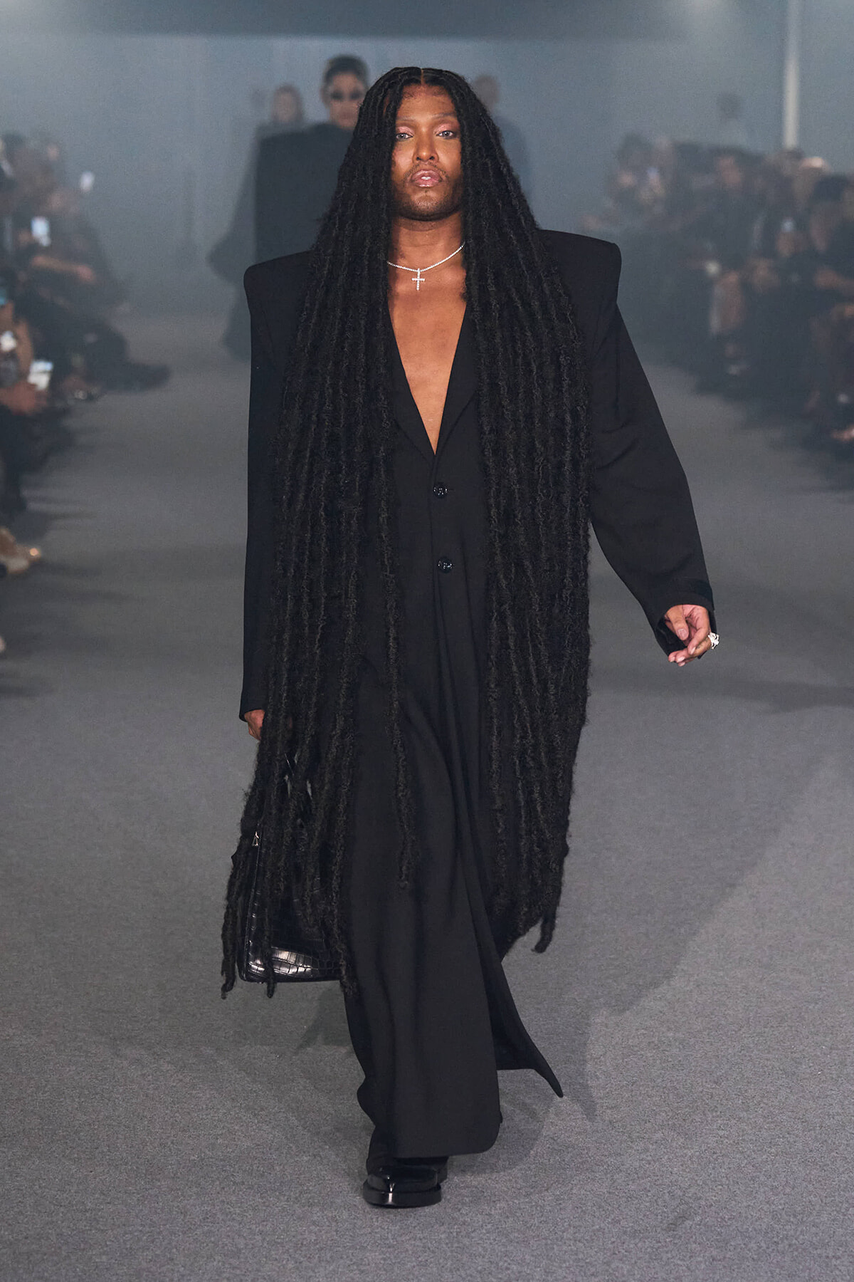 Model walks a runway in a loose black suit with extreme long dreadlocks and a cross necklace, minimal makeup.