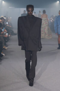 Model walking a fashion runway in an oversized black double-breasted blazer and matching pants, wearing sunglasses.