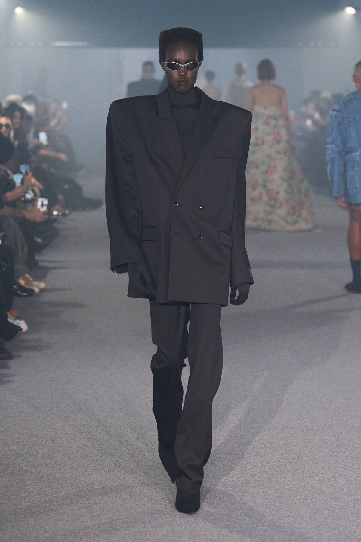 Model walking a fashion runway in an oversized black double-breasted blazer and matching pants, wearing sunglasses.