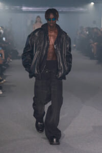 Male model on runway wearing an oversized black leather jacket, hoodie, baggy black pants, and blue sunglasses.