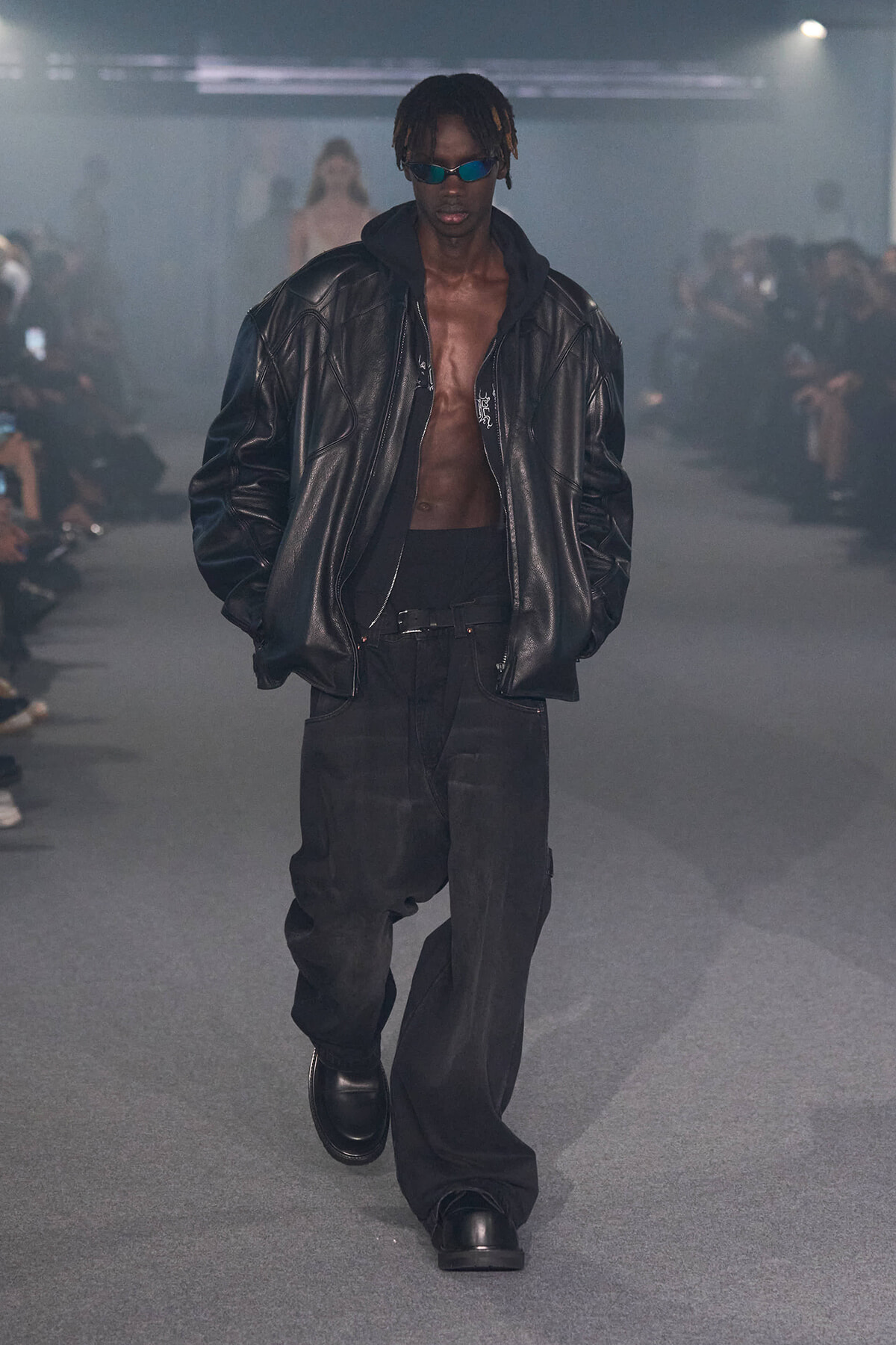 Male model on runway wearing an oversized black leather jacket, hoodie, baggy black pants, and blue sunglasses.