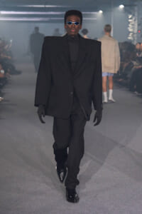 Male model in an oversized black suit and turtleneck walks a runway, wearing dark sunglasses and gloves for a dramatic, monochrome look.