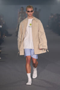 Male model walking a fashion runway in a tan oversized jacket, white graphic tee, light blue striped shorts, white socks, and sunglasses.