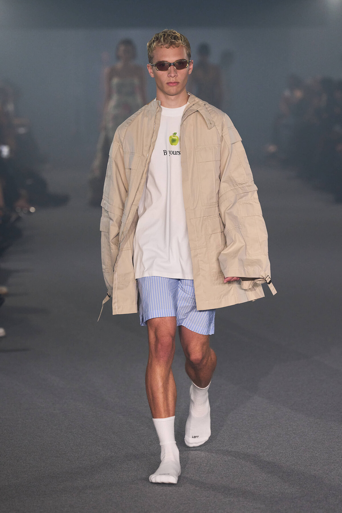 Male model walking a fashion runway in a tan oversized jacket, white graphic tee, light blue striped shorts, white socks, and sunglasses.