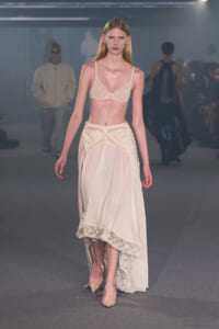 Model walking a fashion runway in cream lace lingerie and a sheer, lace-trim skirt.