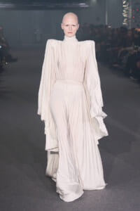Bald model wearing a flowing ivory pleated gown with dramatic cape sleeves on a runway.