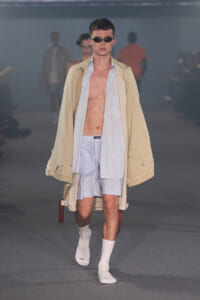 Male model walks the runway in an oversized beige trench over a pale blue shirt and light blue shorts, with white socks and sunglasses.