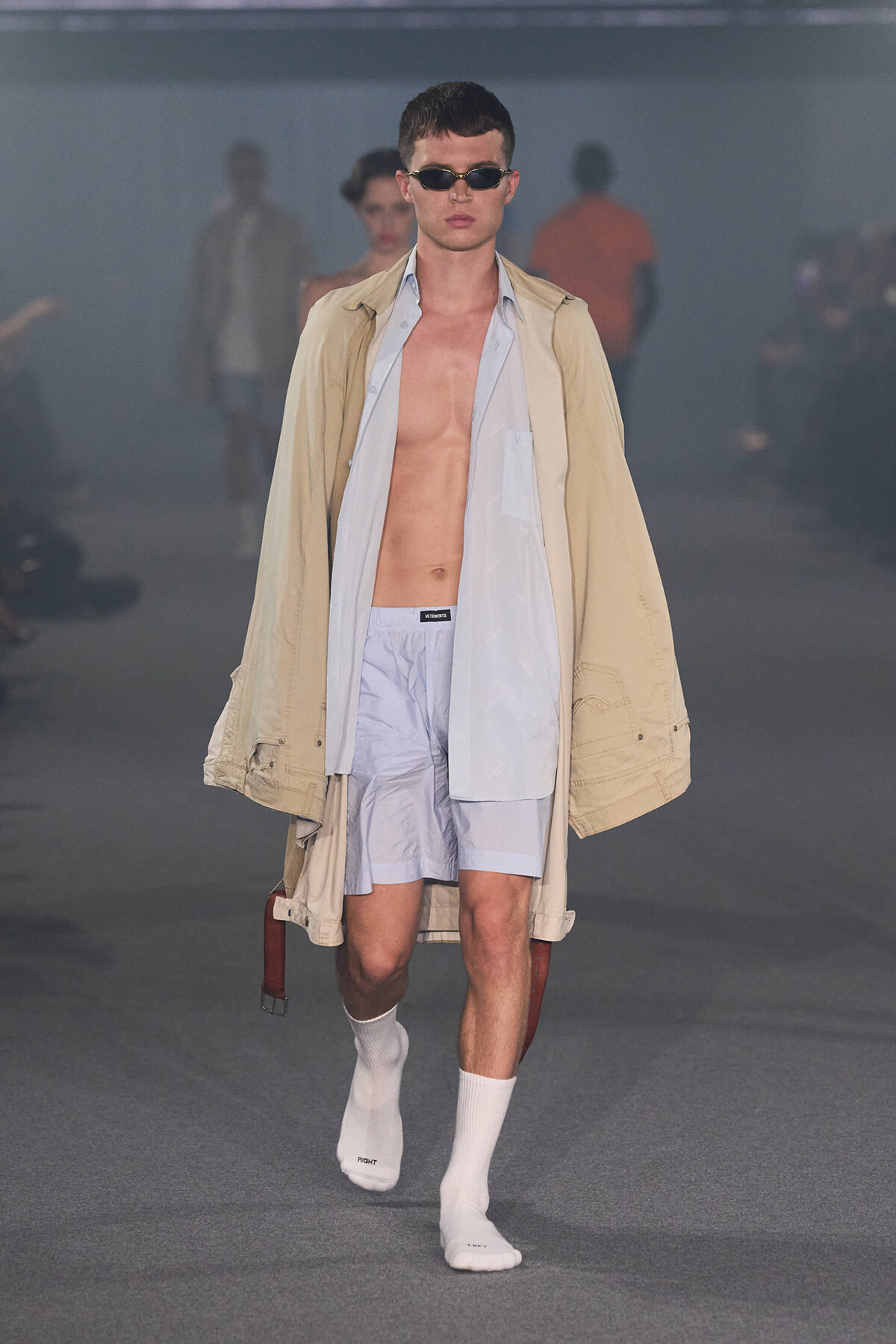Male model walks the runway in an oversized beige trench over a pale blue shirt and light blue shorts, with white socks and sunglasses.