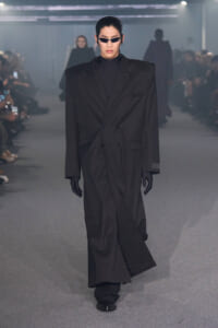 Model walks the runway in an oversized black double-breasted coat, black turtleneck, gloves, and slim sunglasses.
