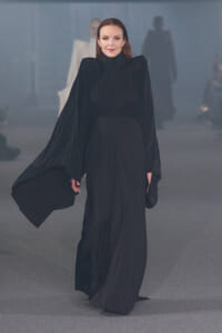 Model walks the runway in a floor-length black pleated gown with cape-like sleeves and a high neckline.