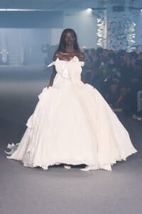 Black model wearing an off-shoulder white wedding gown with a long train walking a runway.