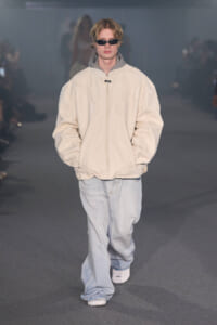 Male model walking a runway in an oversized beige padded jacket, light gray hood, and baggy light-wash jeans with sunglasses.