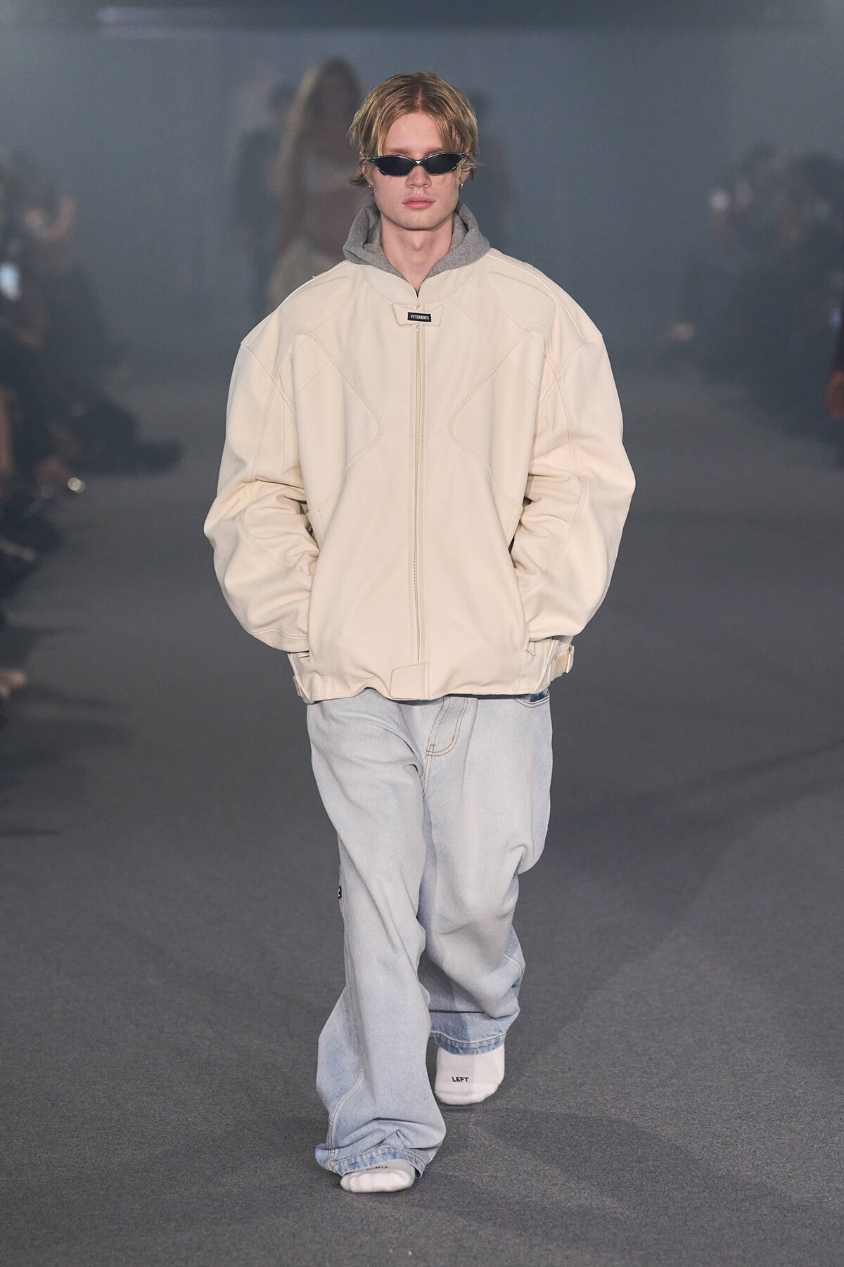 Male model walking a runway in an oversized beige padded jacket, light gray hood, and baggy light-wash jeans with sunglasses.