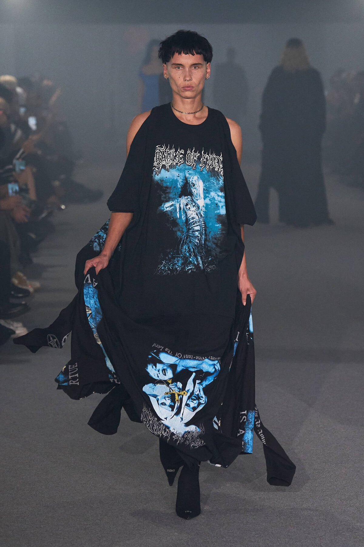 Models walks a fashion runway in an oversized black outfit with blue graphic print on the chest and a large blue graphic at the hem.