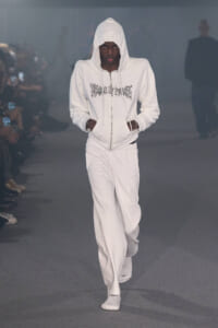 Male model in an all-white hoodie and track pants walks a fashion runway, hands in pockets, hood up.