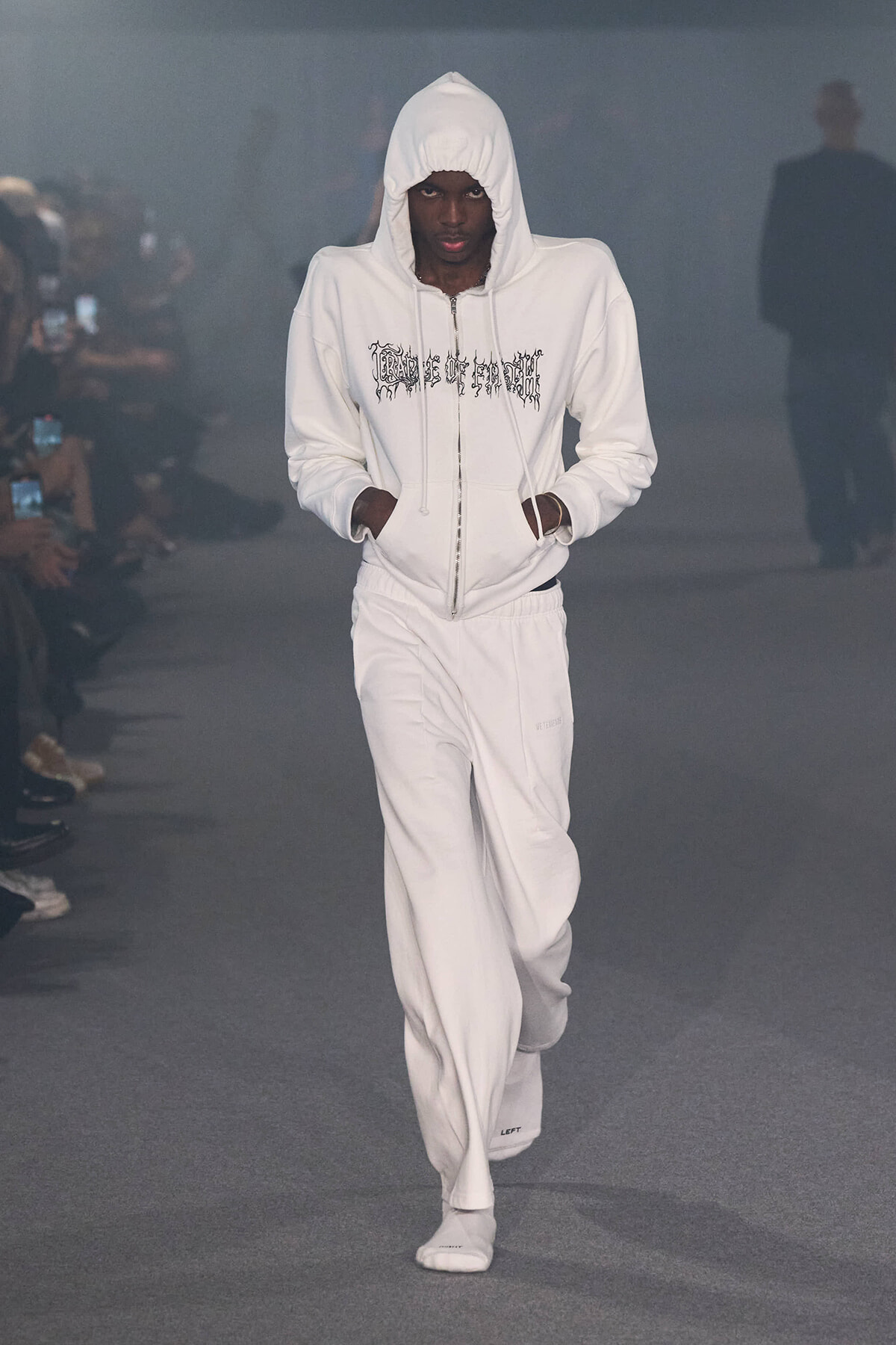 Male model in an all-white hoodie and track pants walks a fashion runway, hands in pockets, hood up.