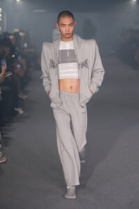 Male model on a runway wearing a light gray tracksuit with a cropped striped top and hands in pockets.