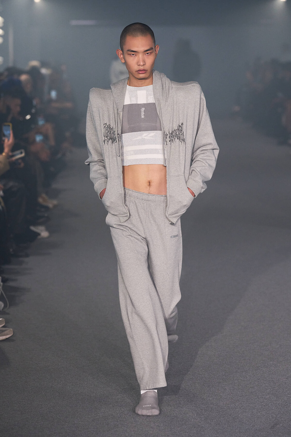 Male model on a runway wearing a light gray tracksuit with a cropped striped top and hands in pockets.