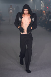 Male fashion model on a runway in a black zip hoodie opened to show chest tattoos, with black pants and socks.