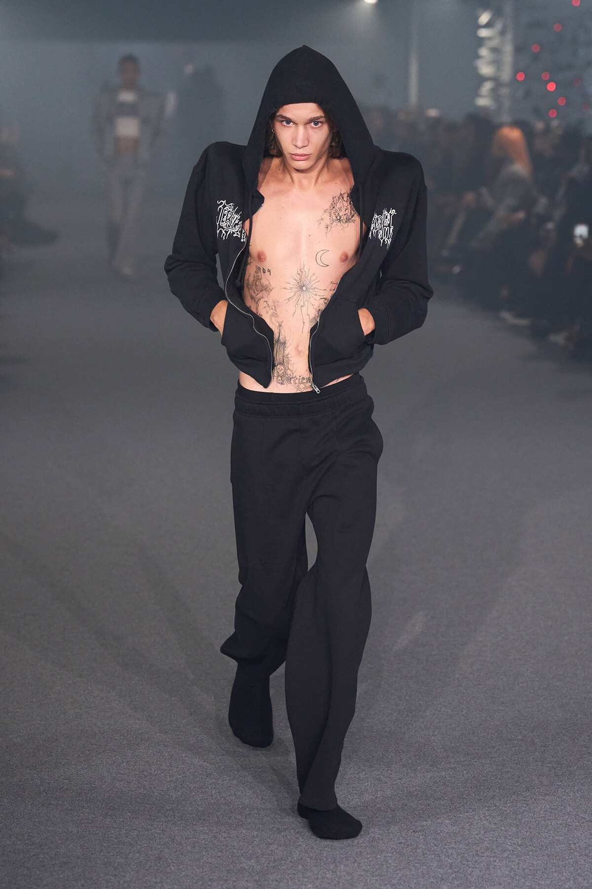 Male fashion model on a runway in a black zip hoodie opened to show chest tattoos, with black pants and socks.