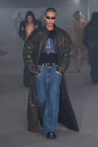 Male model in oversized black leather trench and wide jeans walks a fashion runway, wearing a Paris-themed graphic tee and dark sunglasses.