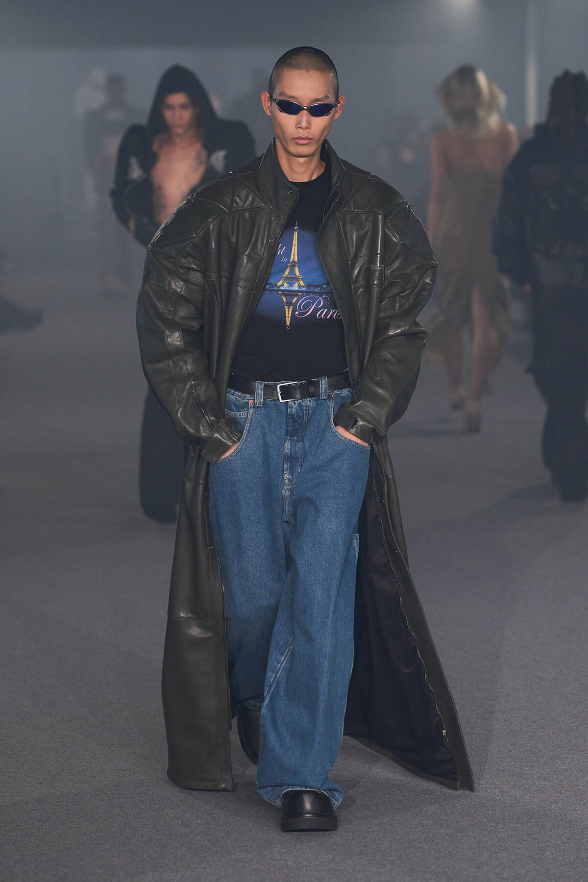 Male model in oversized black leather trench and wide jeans walks a fashion runway, wearing a Paris-themed graphic tee and dark sunglasses.