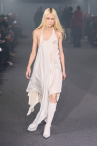 Model walks the runway in a sheer, flowing white dress with lace trim and an asymmetrical hem, paired with white knee-high boots in a gray backstage-lit setting.