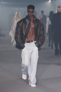 Male model on a runway wearing a dark brown oversized leather jacket, open to reveal his bare chest, light baggy jeans, and white sneakers, with sunglasses.