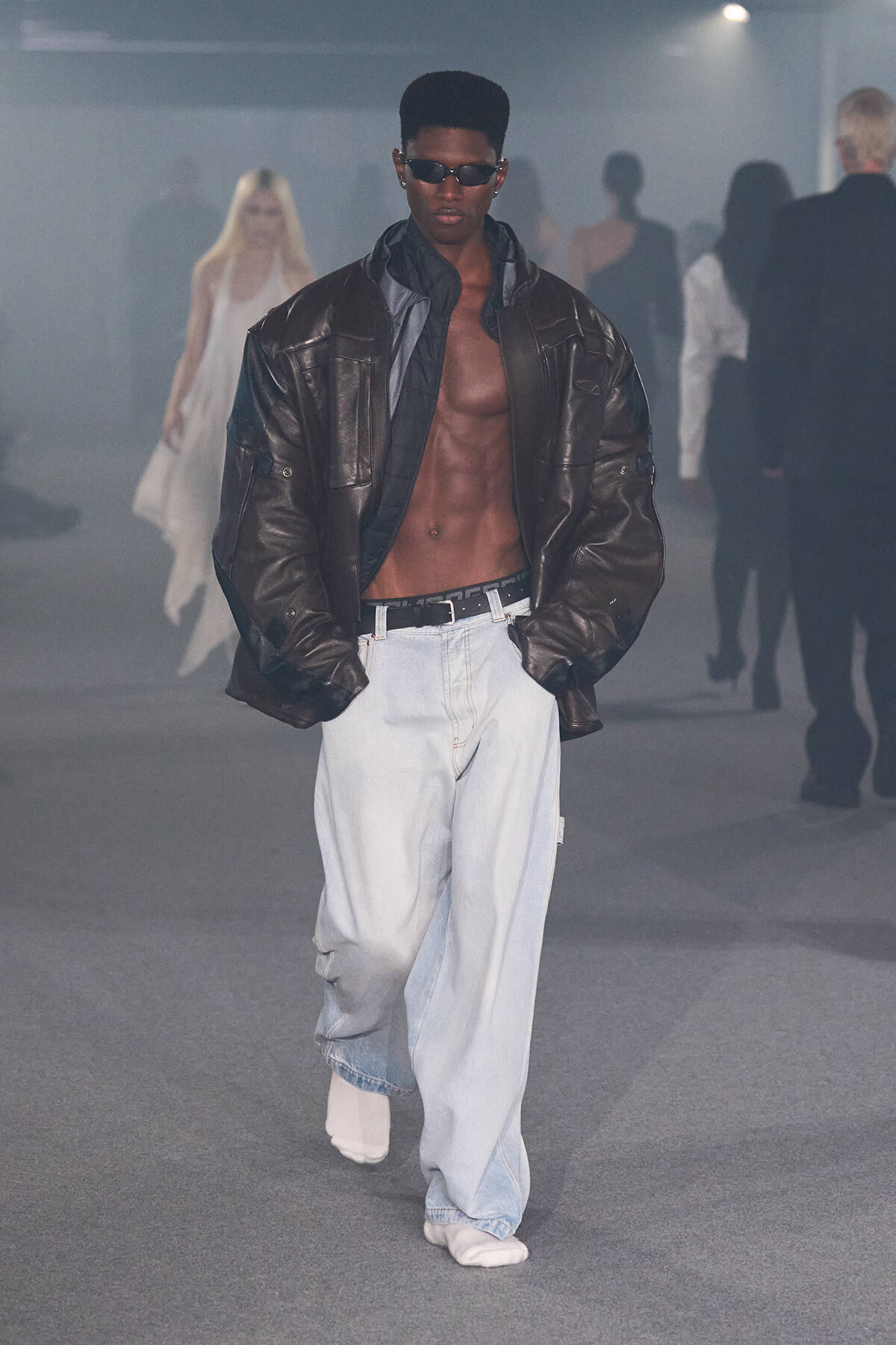 Male model on a runway wearing a dark brown oversized leather jacket, open to reveal his bare chest, light baggy jeans, and white sneakers, with sunglasses.