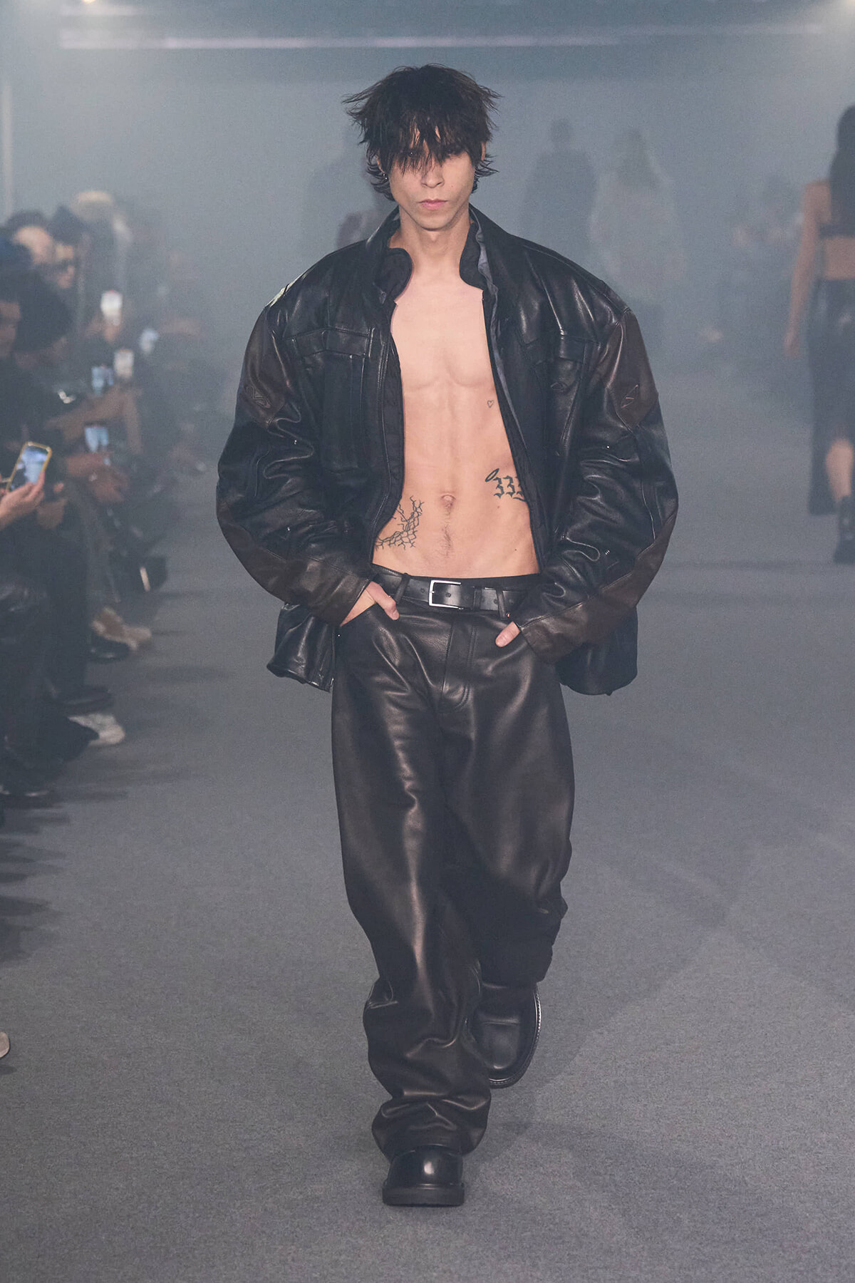 Male model walking the runway in a black leather jacket and pants, chest exposed with visible tattoos.