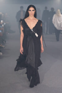 Model walking the runway in a black, flowing gown with ruffled lace details and a white tag on the dress.